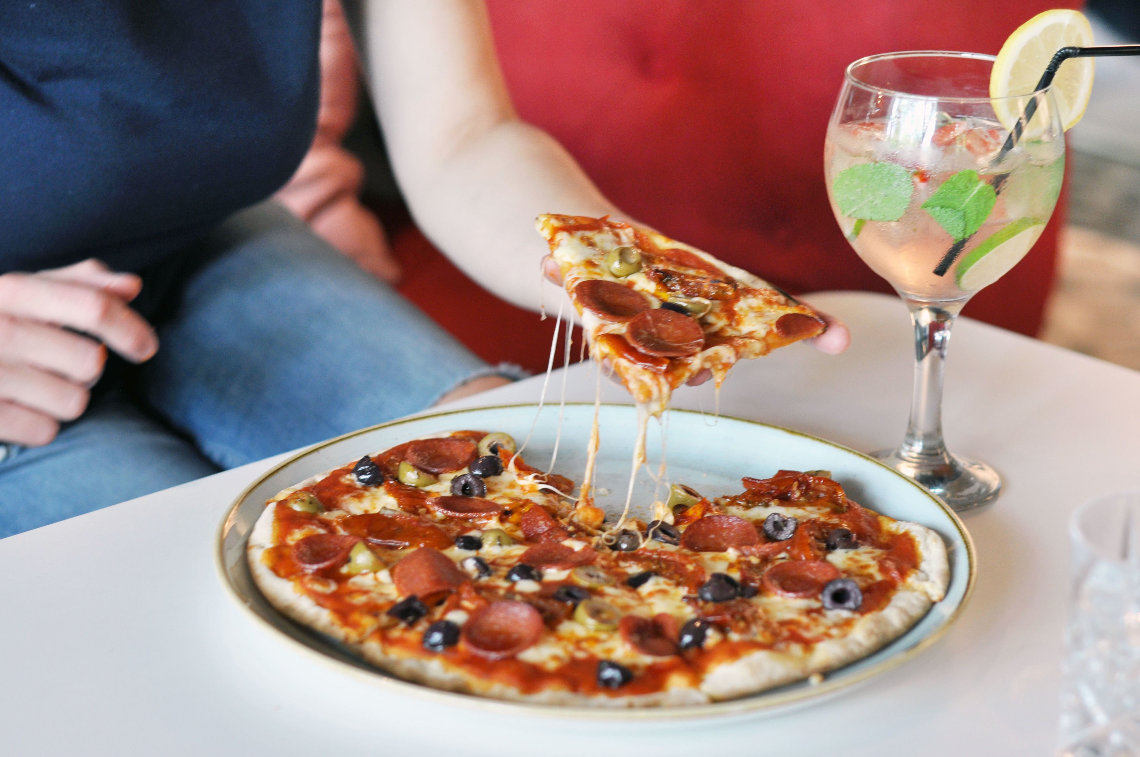 Une pizza garnie de pepperoni et d’olives, présentée sur une assiette, un verre contenant une boisson avec une paille, et une part de pizza en train d’être soulevée.