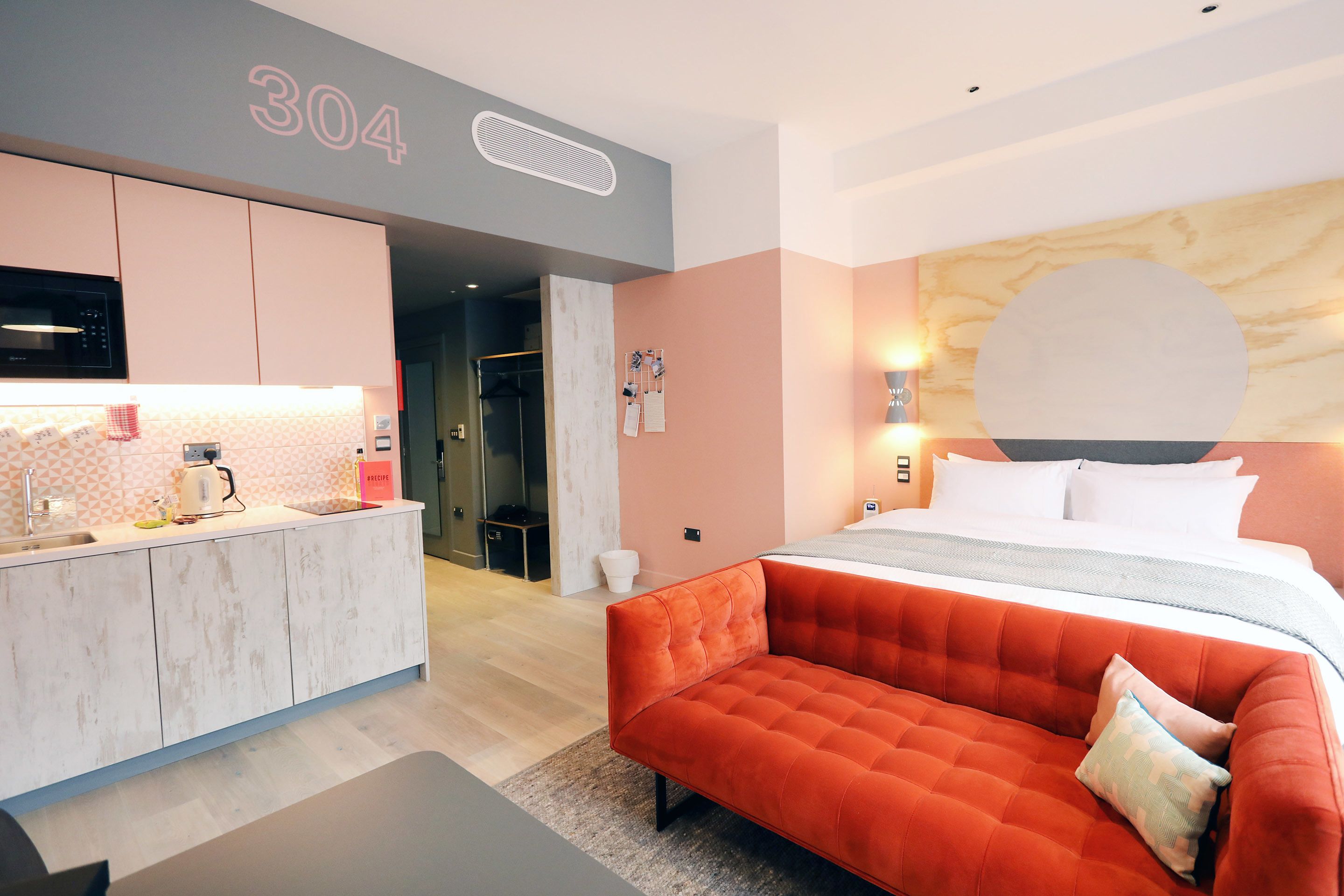 A modern hotel room with a kitchenette, a large bed with a headboard, and a red sofa.