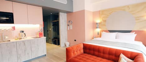 A modern hotel room with a kitchenette, a large bed with a headboard, and a red sofa.