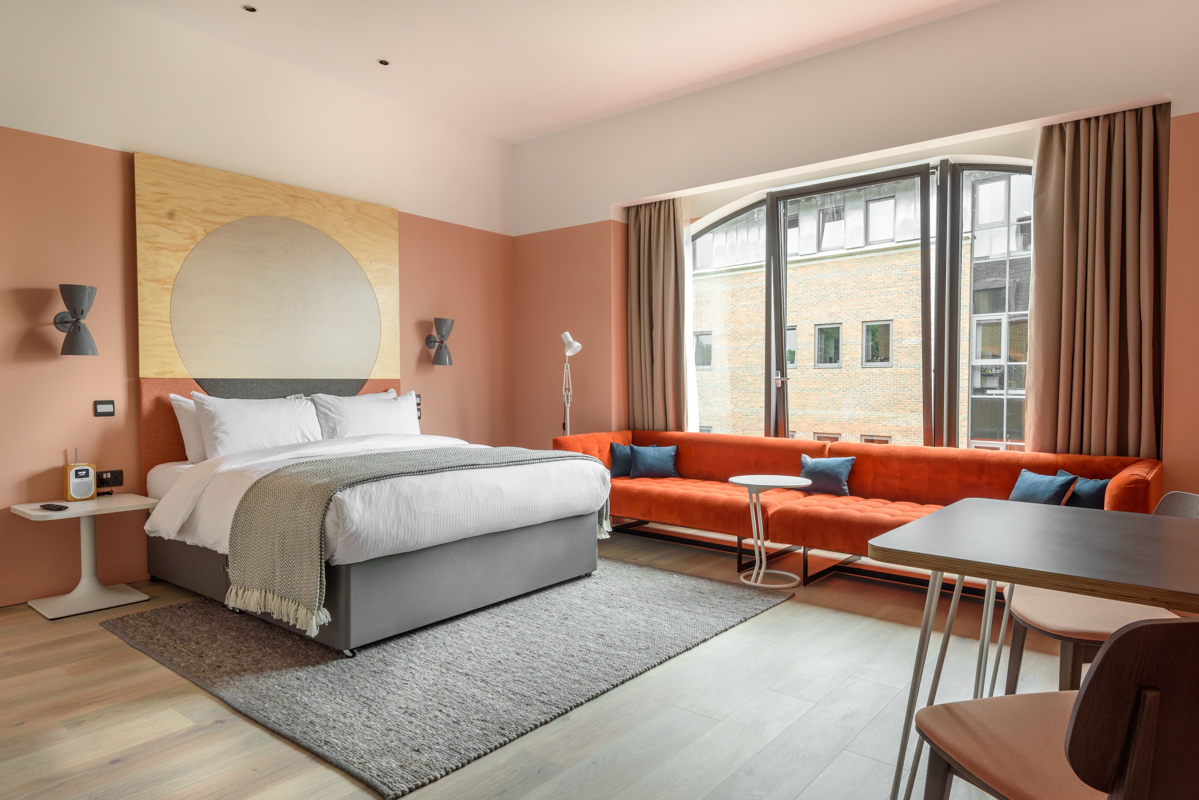 A modern hotel room with a large bed, an orange sofa, a small table, and a view of a brick building through large windows.
