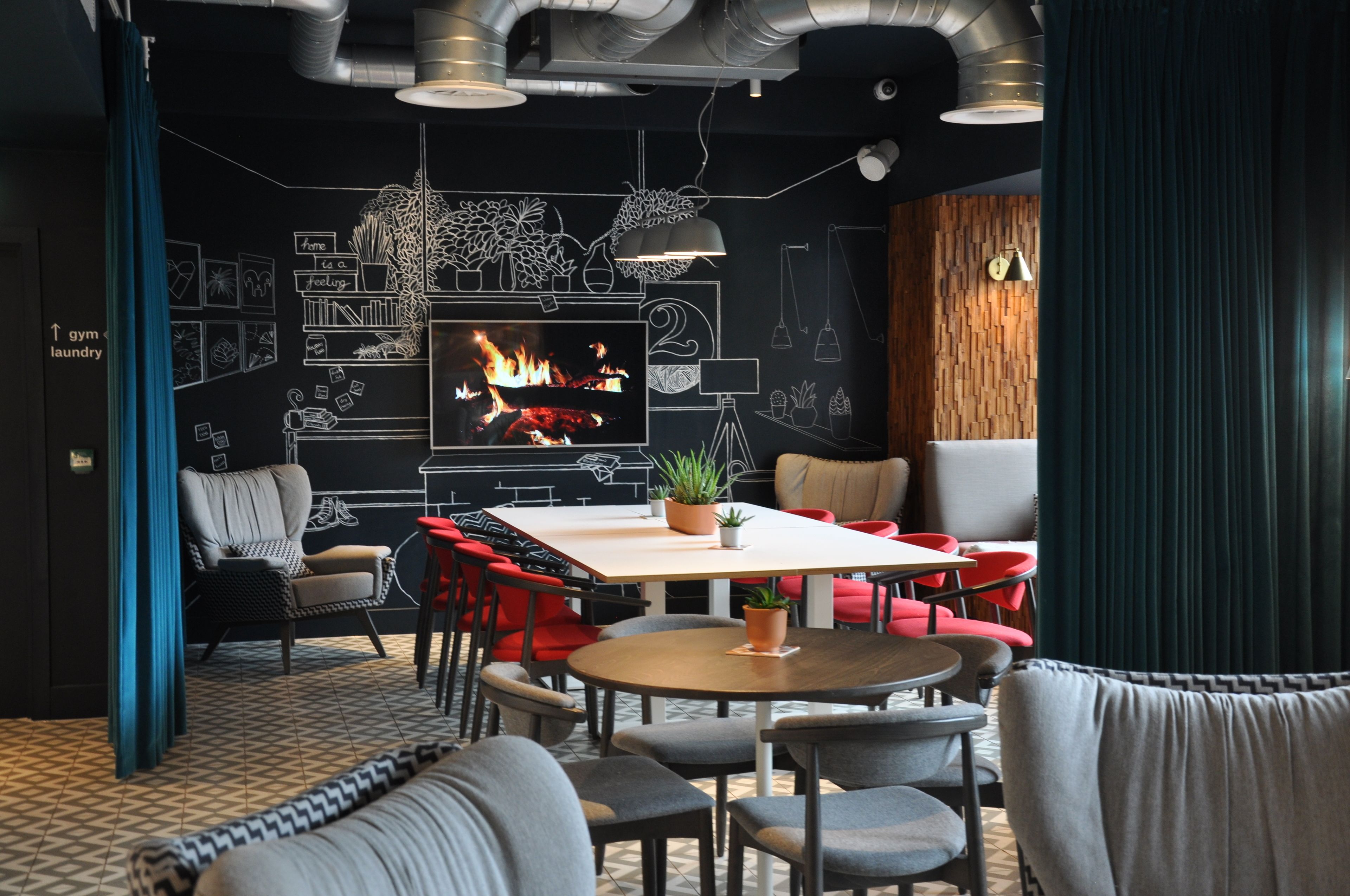 A modern lounge area with a large flat-screen TV displaying a fire, a wooden wall with chalkboard drawings, and a variety of seating options including red chairs and a sofa.