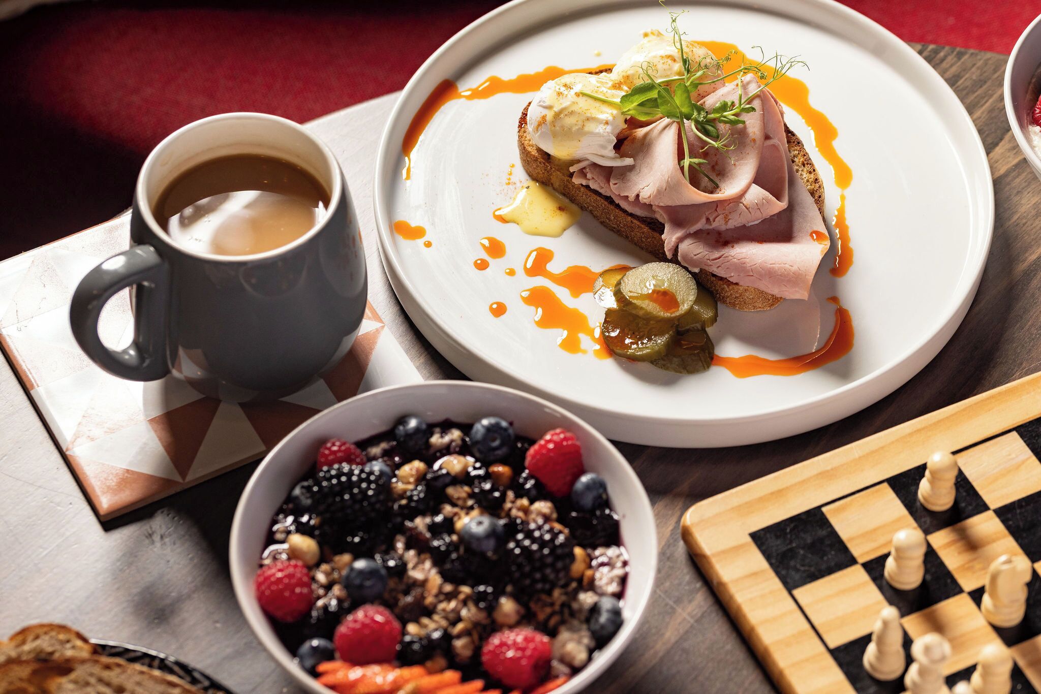 Mesa de café da manhã com prato de comida, xícara de café, tigela de frutas e um tabuleiro de xadrez.