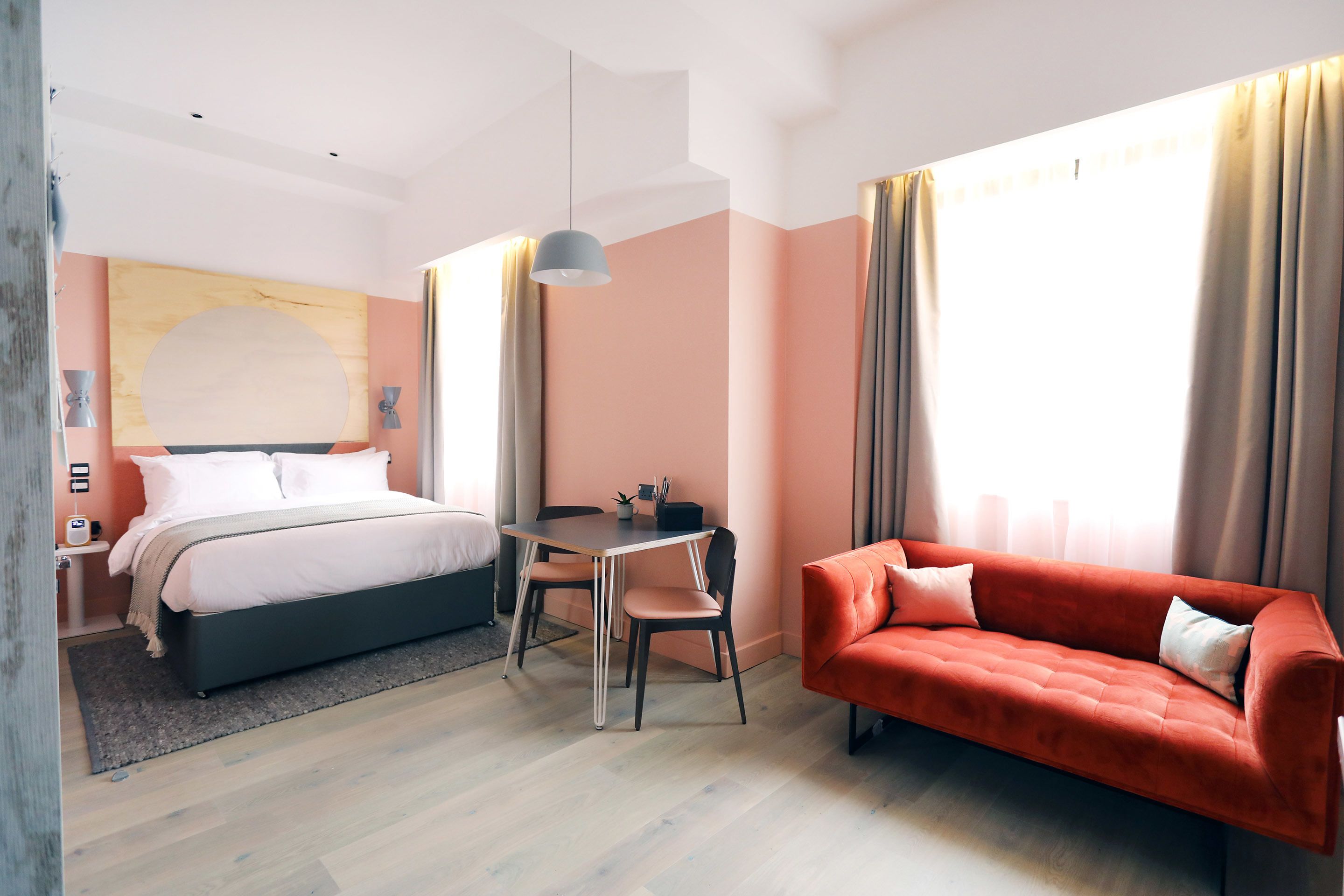 Een moderne hotelkamer met een bed, een rode bank, een eettafel en een groot raam met gordijnen.
