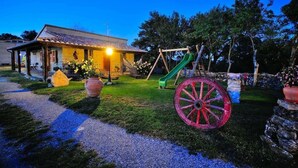 Uma casa com varanda, baloiço e uma grande roda de carroça vermelha no jardim.