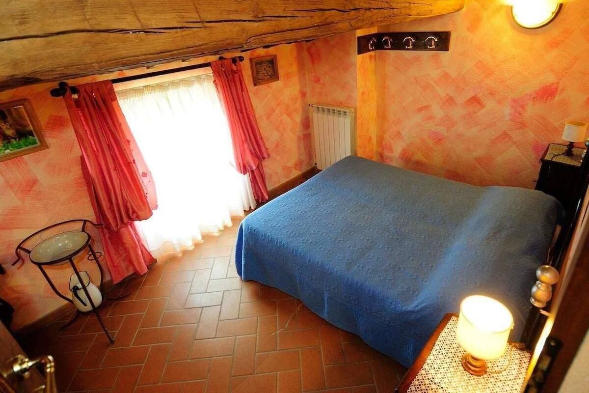 Een slaapkamer met een bed, een klein tafeltje met een lamp, een radiator en een raam met gordijnen.
