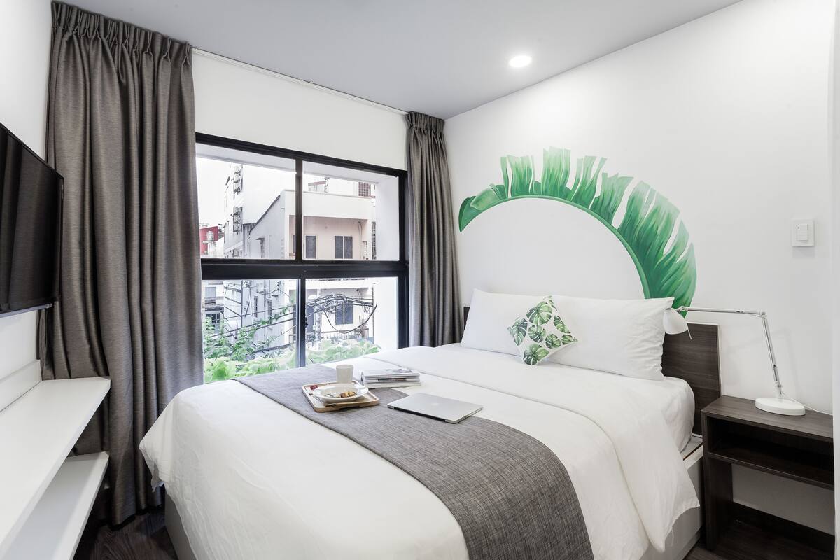 Quarto de hotel com cama, escrivaninha, janela com cortinas, luminária na parede e decoração com folha verde.