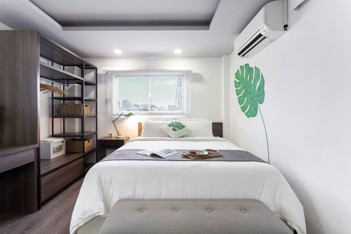 Quarto moderno com cama, guarda-roupa, janela com vista para a cidade e uma grande folha verde decorativa na parede.