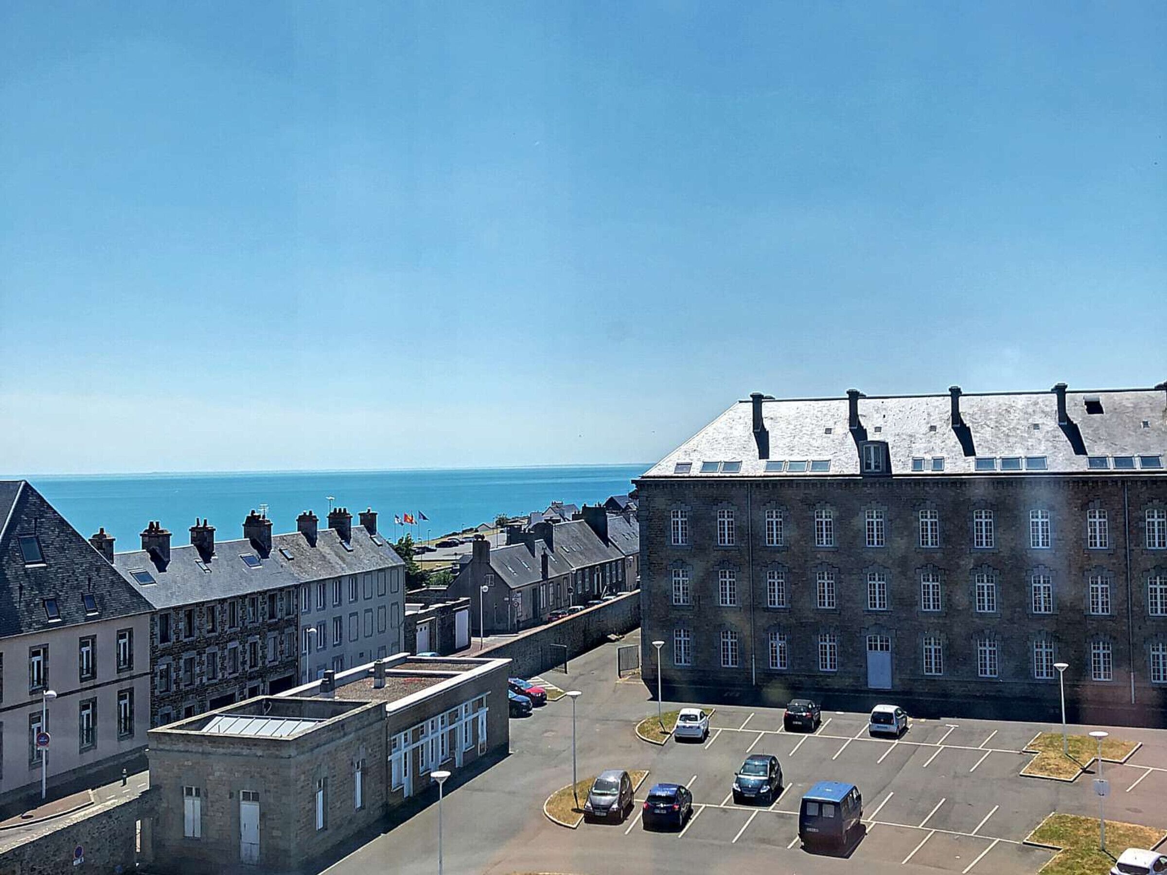 A parking lot with several cars, a large building with multiple windows, and a clear view of the sea in the background.