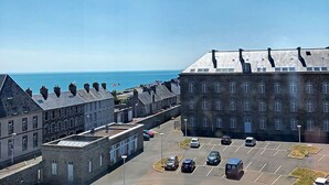 Un parking avec plusieurs voitures, un grand bâtiment doté de nombreuses fenêtres et, au loin, une vue dégagée sur la mer.