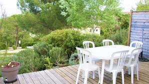 Une terrasse en bois avec une table et des chaises blanches, entourée de verdure et d’arbres.