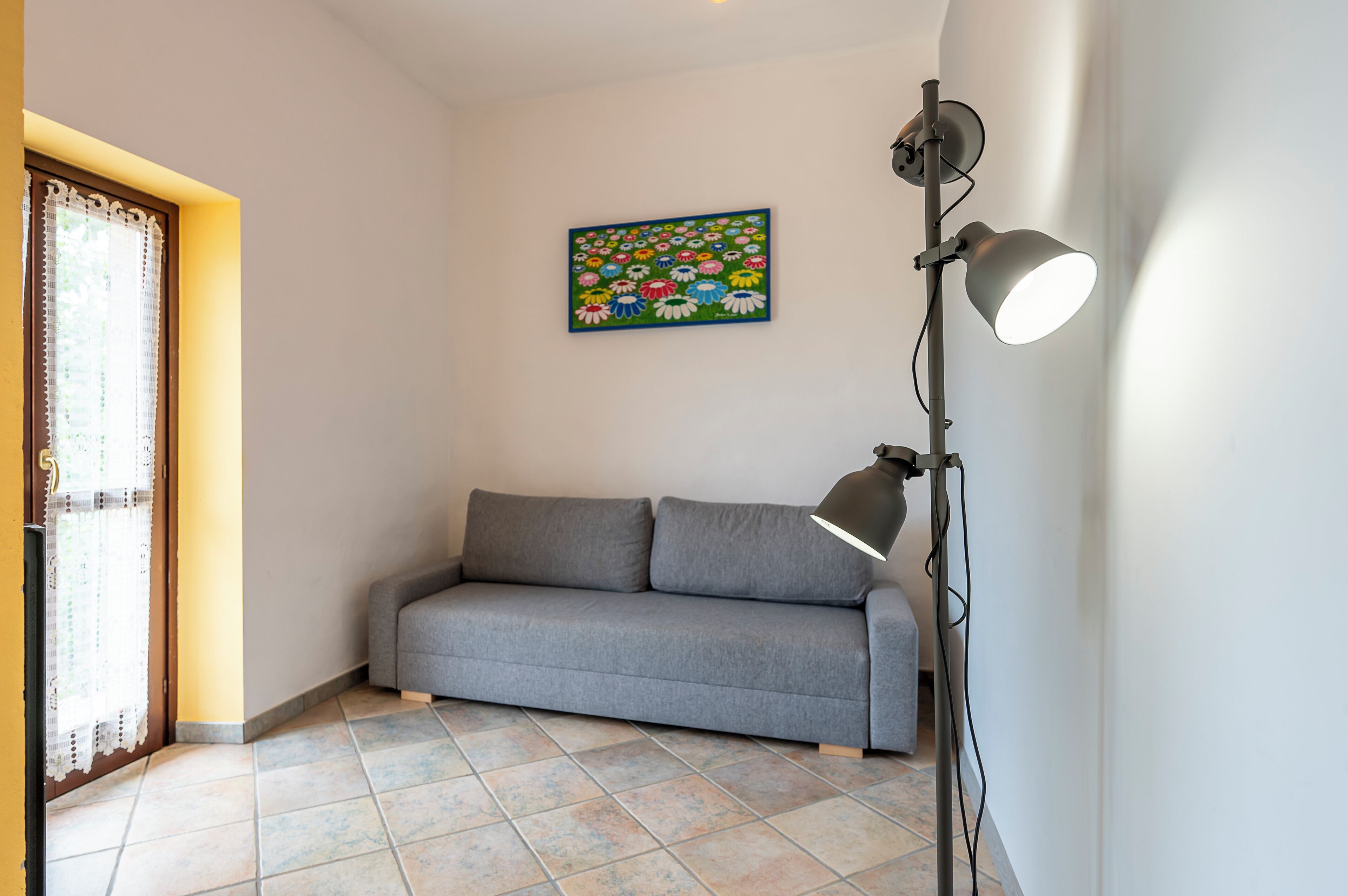 A room with a grey sofa, a floor lamp, and a colorful floral painting on the wall.