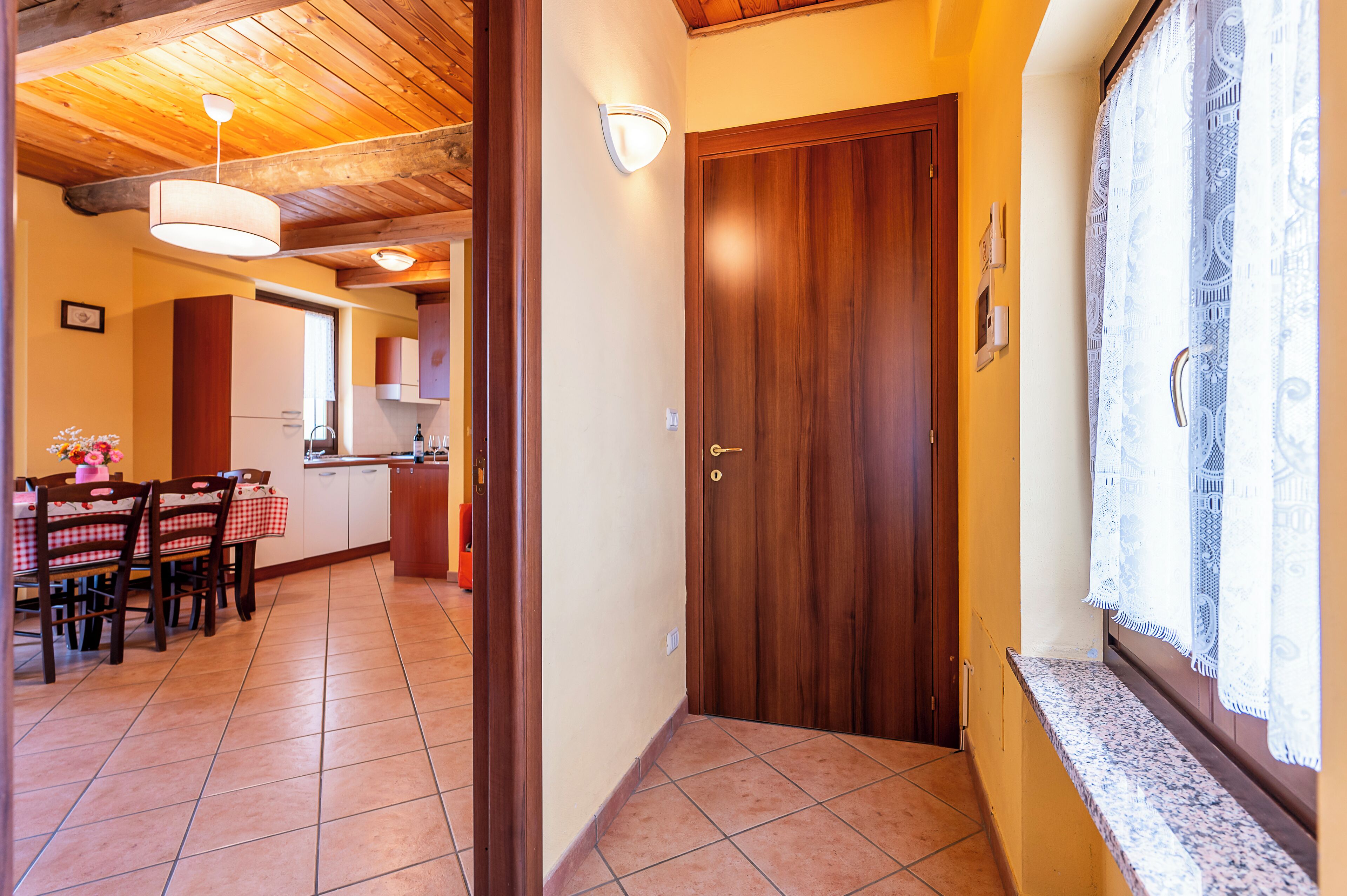 Un couloir avec une porte en bois, une fenêtre ornée de rideaux en dentelle et un coin cuisine avec une table et des chaises.