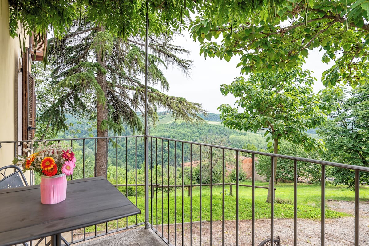 Een balkon met een tafel en een roze vaas met bloemen, met uitzicht op een weelderig groen landschap.