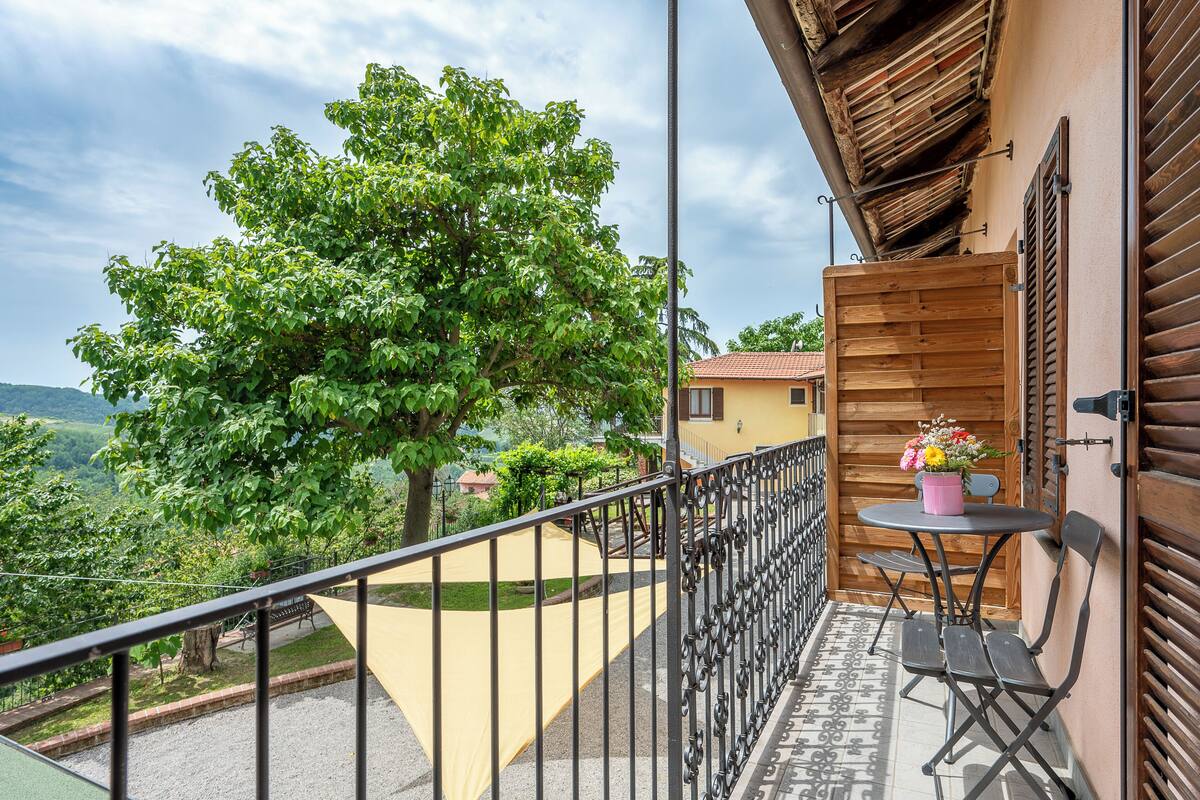 Een balkon met uitzicht op een boom en heuvels, een tafel met stoelen en een houten deur.