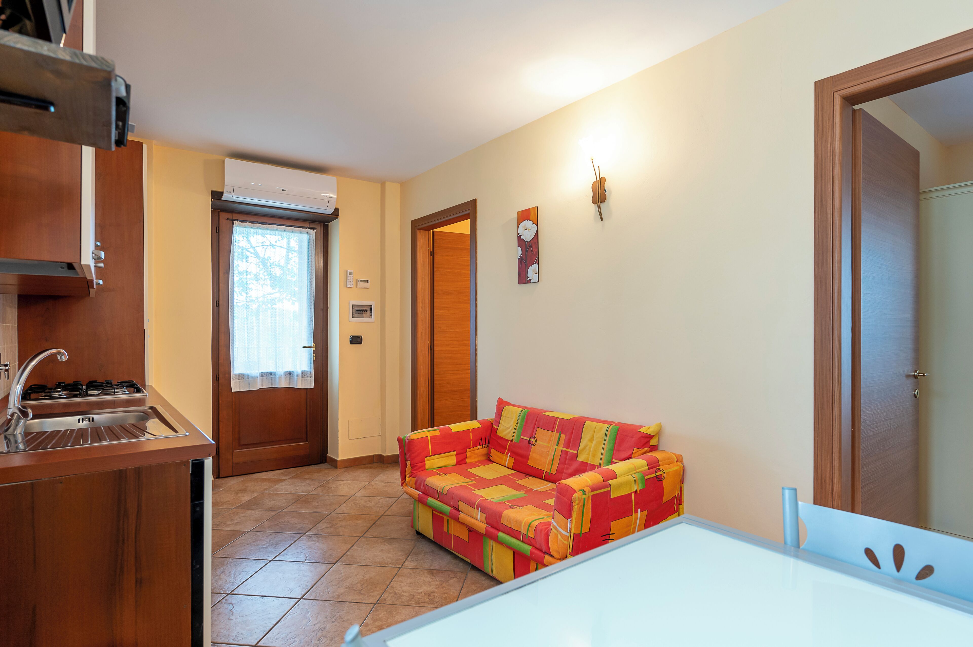 Una cocina compacta con estufa y lavamanos integrados, una puerta de madera que da a otra habitación, y un sofá colorido en una pequeña sala de estar.