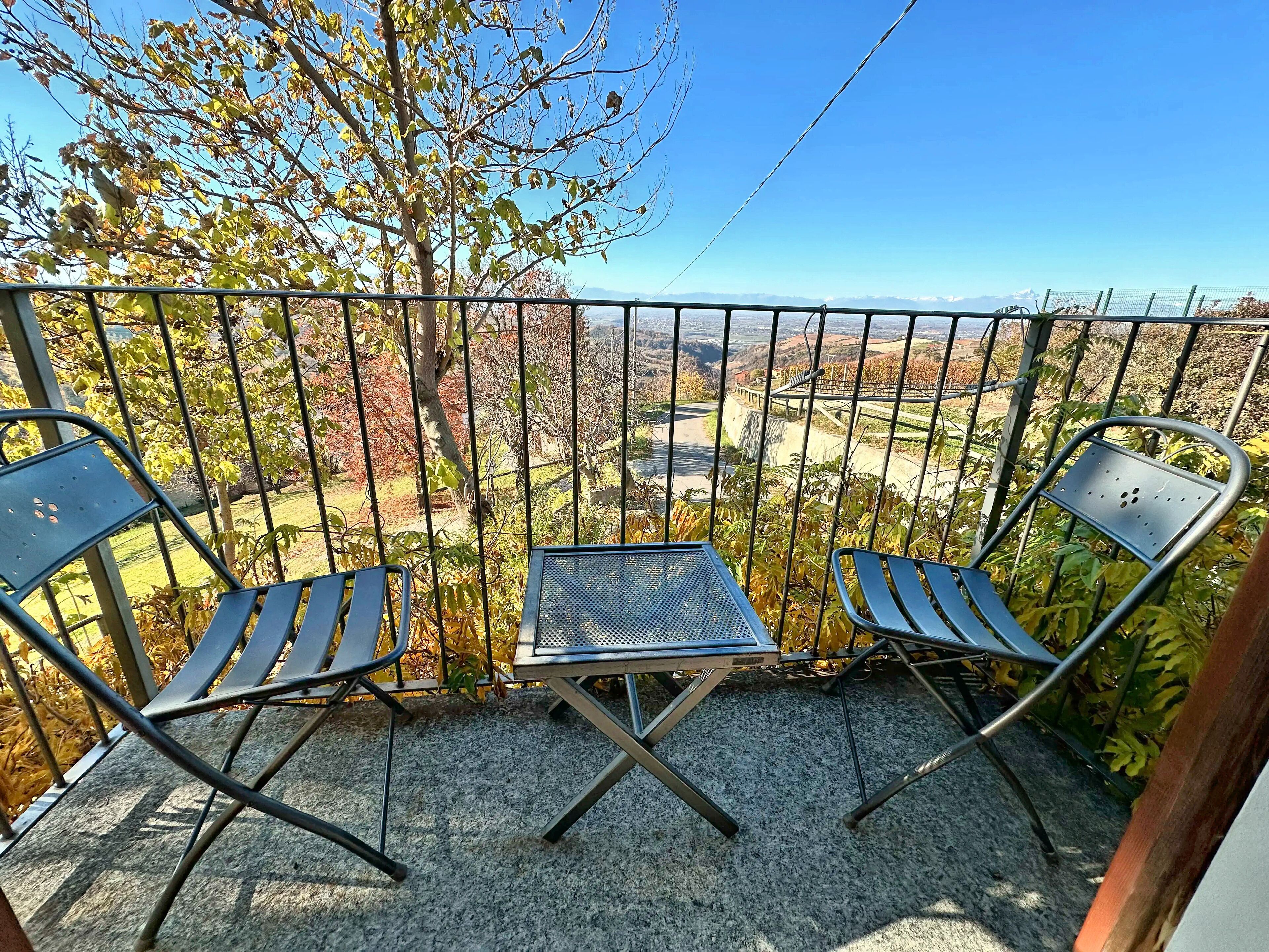 A balcony with two metal chairs and a small table, overlooking a scenic landscape with trees and a distant view.