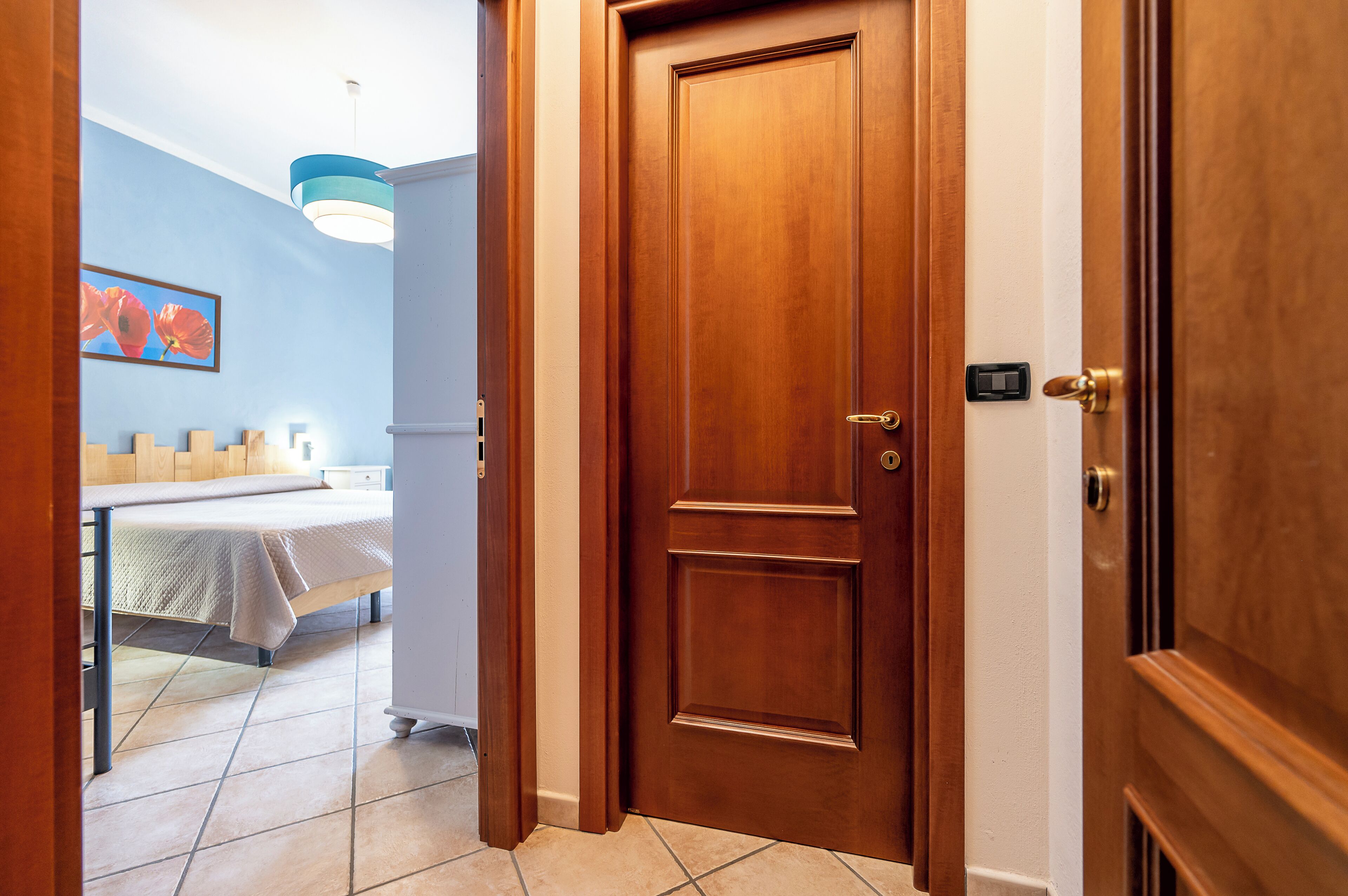 Habitación de hotel con puerta de madera, cama con cubrecama blanco, mesita de noche y pared azul con un cuadro enmarcado.