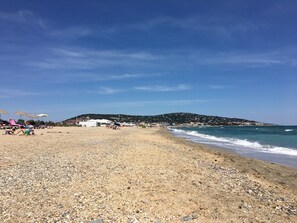 A pebble beach with umbrellas and beach chairs, a hill in the background, and a clear sky.