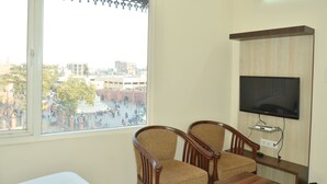 A room with a large window, two wooden chairs, a flat-screen TV mounted on the wall, and a small table. - Hotel Temple Inn (Amritsar)