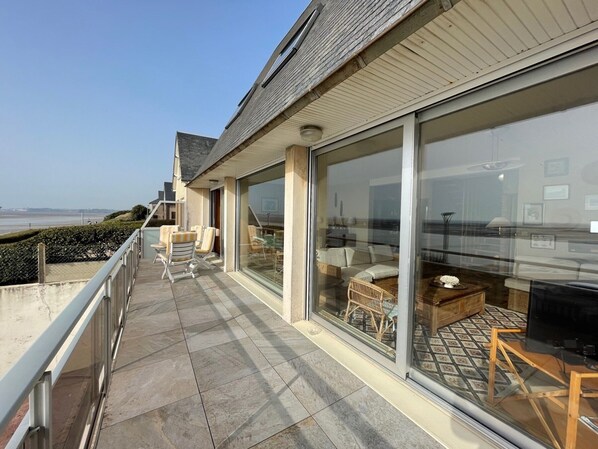A balcony with a view of the sea, furnished with chairs and a table, and a glass door leading to an interior space with a sofa and a TV. - Seafront villa with garden and free Wifi (Saint-Pair-sur-Mer)
