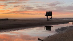 Uma praia com uma torre salva-vidas, águas calmas refletindo o céu e uma faixa de areia.