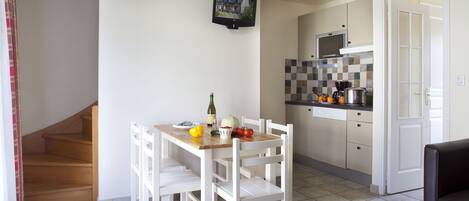 A kitchen with white cabinets, a wooden table, and chairs. There is a microwave, a stove, and a sink. A bottle of wine and some fruits are on the table.