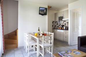 A kitchen with white cabinets, a wooden table, and chairs. There is a microwave, a stove, and a sink. A bottle of wine and some fruits are on the table.