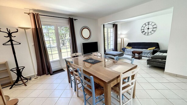 A modern living room with a wooden dining table, blue chairs, a sofa, and a clock on the wall. - Beachside Villa with Garden, Free Wifi & BBQ – Close to Shops & Attractions (Jullouville)