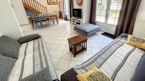 A living room with a sofa, ottoman, and a small table. There is a staircase leading to another room, a dining table with chairs, and a television. - Beachside Villa with Garden, Free Wifi & BBQ – Close to Shops & Attractions (Jullouville)