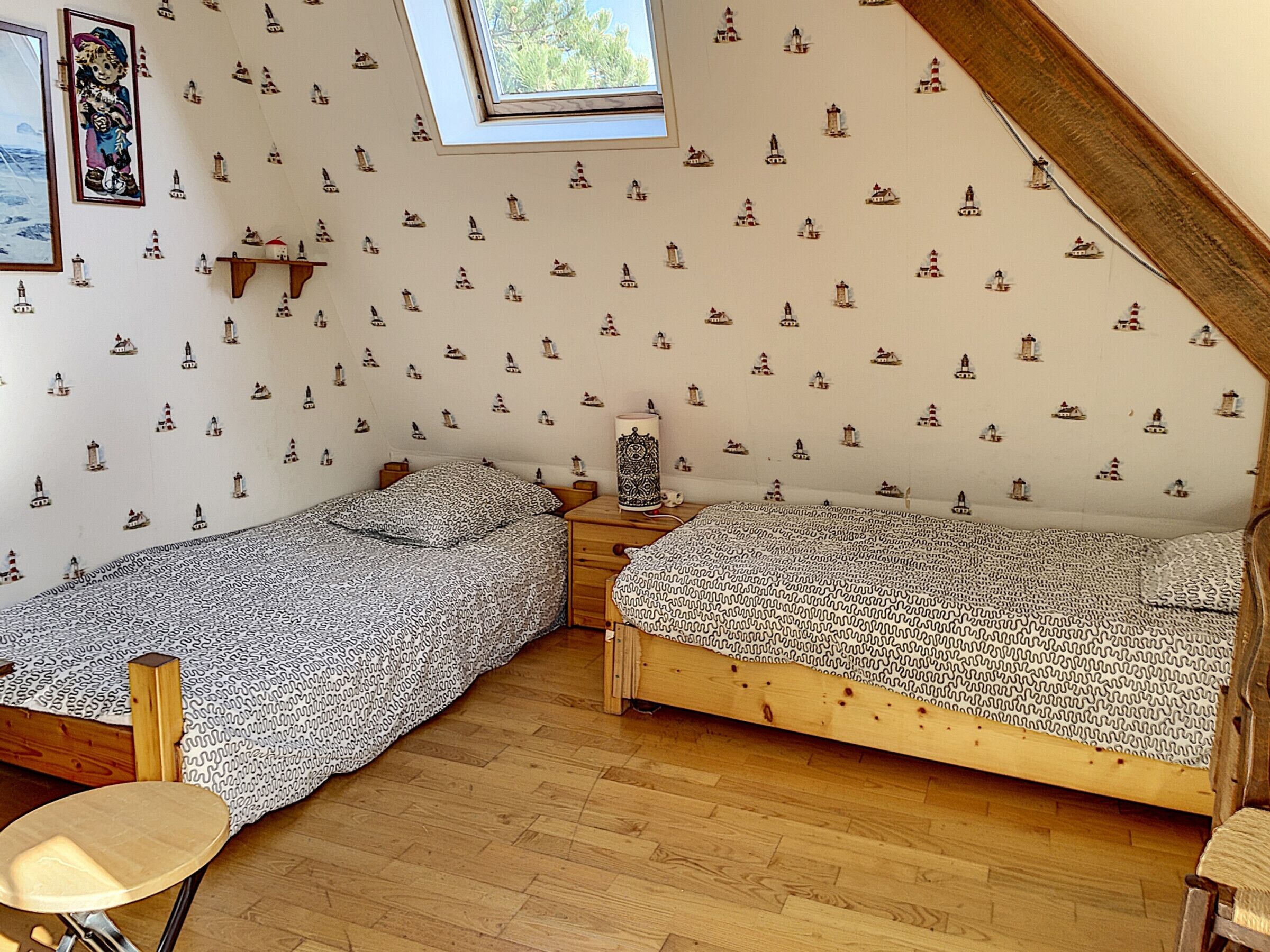 Un dormitorio con dos camas individuales, una mesita, una silla y un tragaluz.