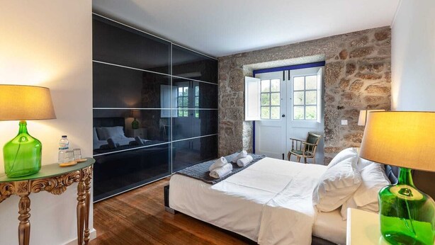 A modern bedroom with a large bed, a glass partition, a stone wall, and a window. - Eido do Carvalhoso (Arcos de Valdevez)