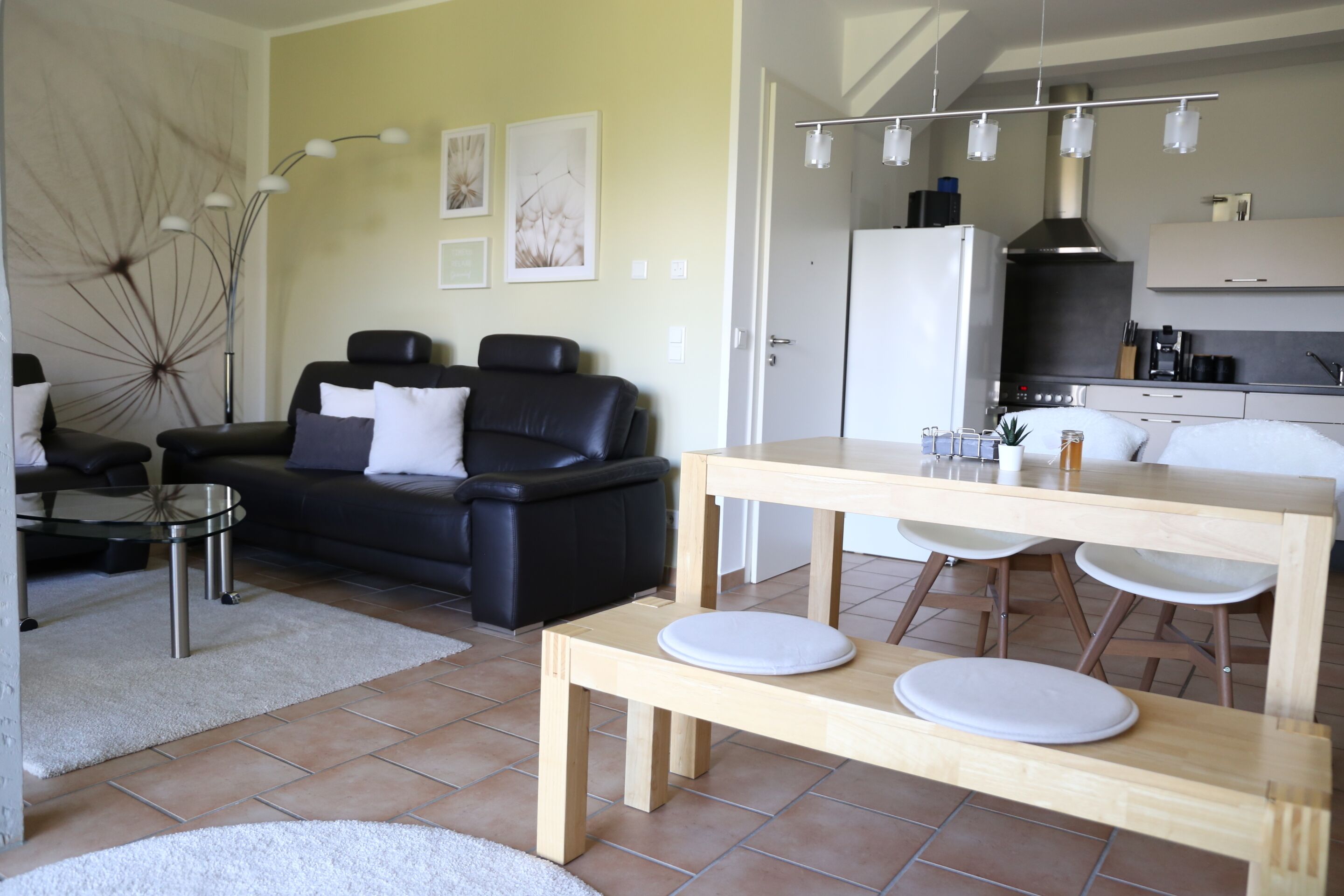 A modern living room with a black leather sofa, a wooden dining table with stools, and a kitchen area with a white refrigerator and stove.