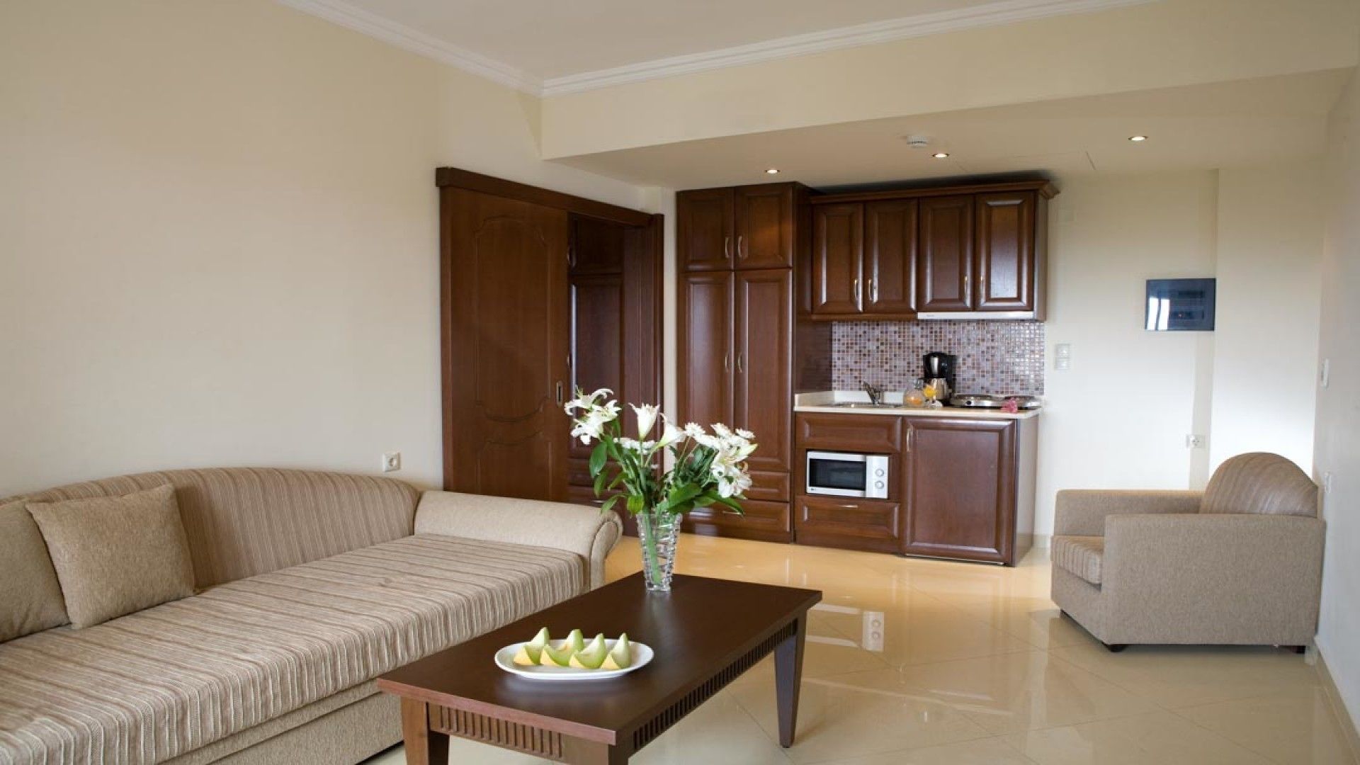 A living room with a sofa, a coffee table with fruit, and a vase of flowers. There is a kitchen area with wooden cabinets and a tiled backsplash.