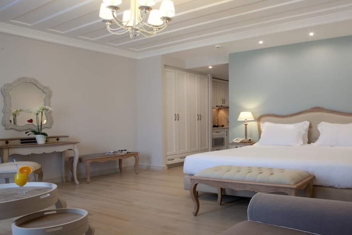 Habitación de hotel con una cama grande, zona de estar con una mesa y sillas, un espejo y una araña.