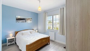 A bedroom with a wooden bed, a nightstand, a lamp, a window with curtains, and a wall-mounted picture. - Cozy Apartment 100m from Beach & Shops, Near Mont Saint-Michel (Jullouville)