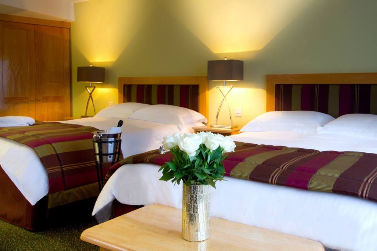 A hotel room with two beds, a wooden headboard, a bedside table with a lamp, and a vase with flowers.