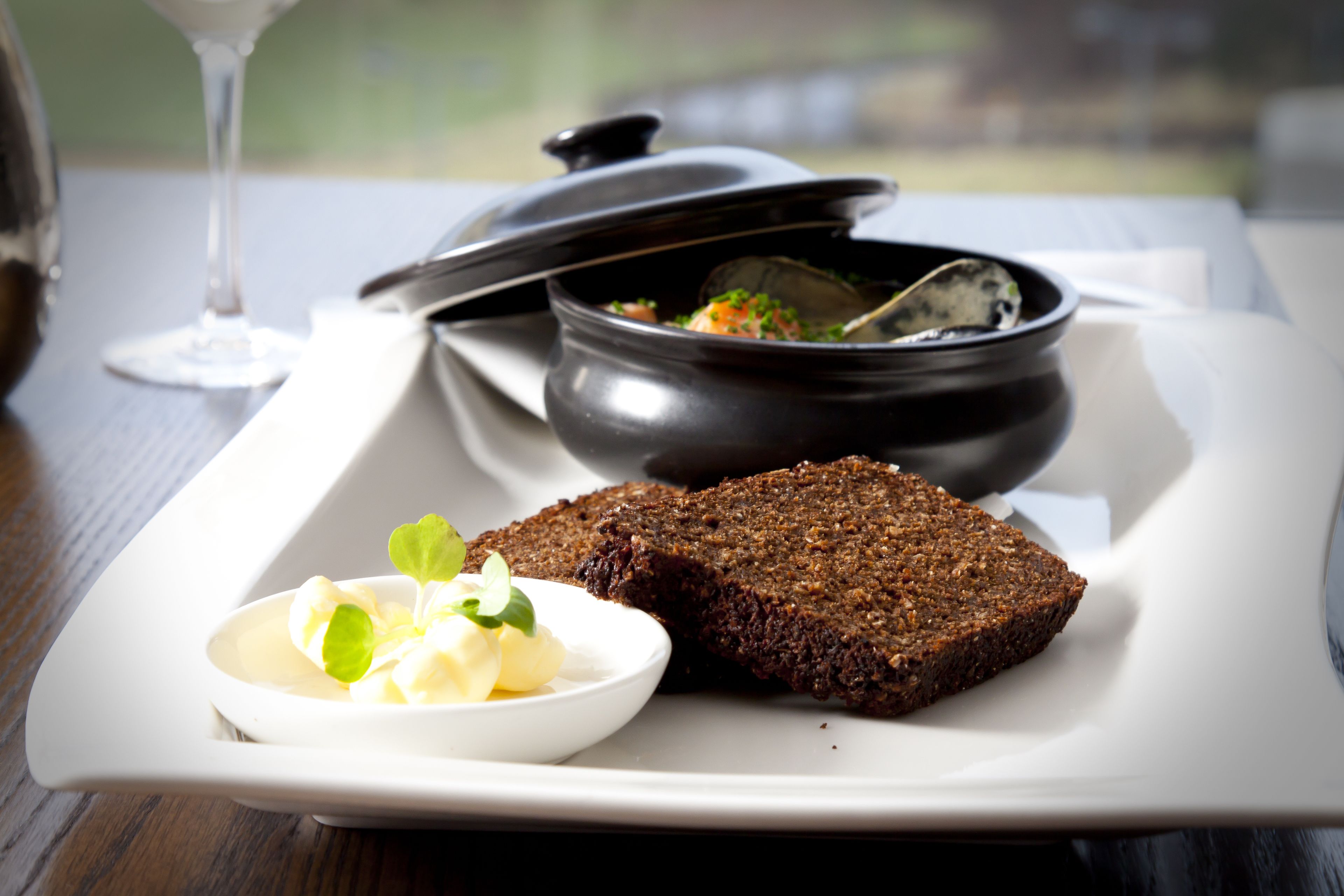 A white plate with a bowl of soup, a piece of dark bread, and a small bowl of butter with herbs.