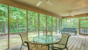 A covered outdoor area with a round glass table and chairs, a bench, and a view of a forest through large windows.