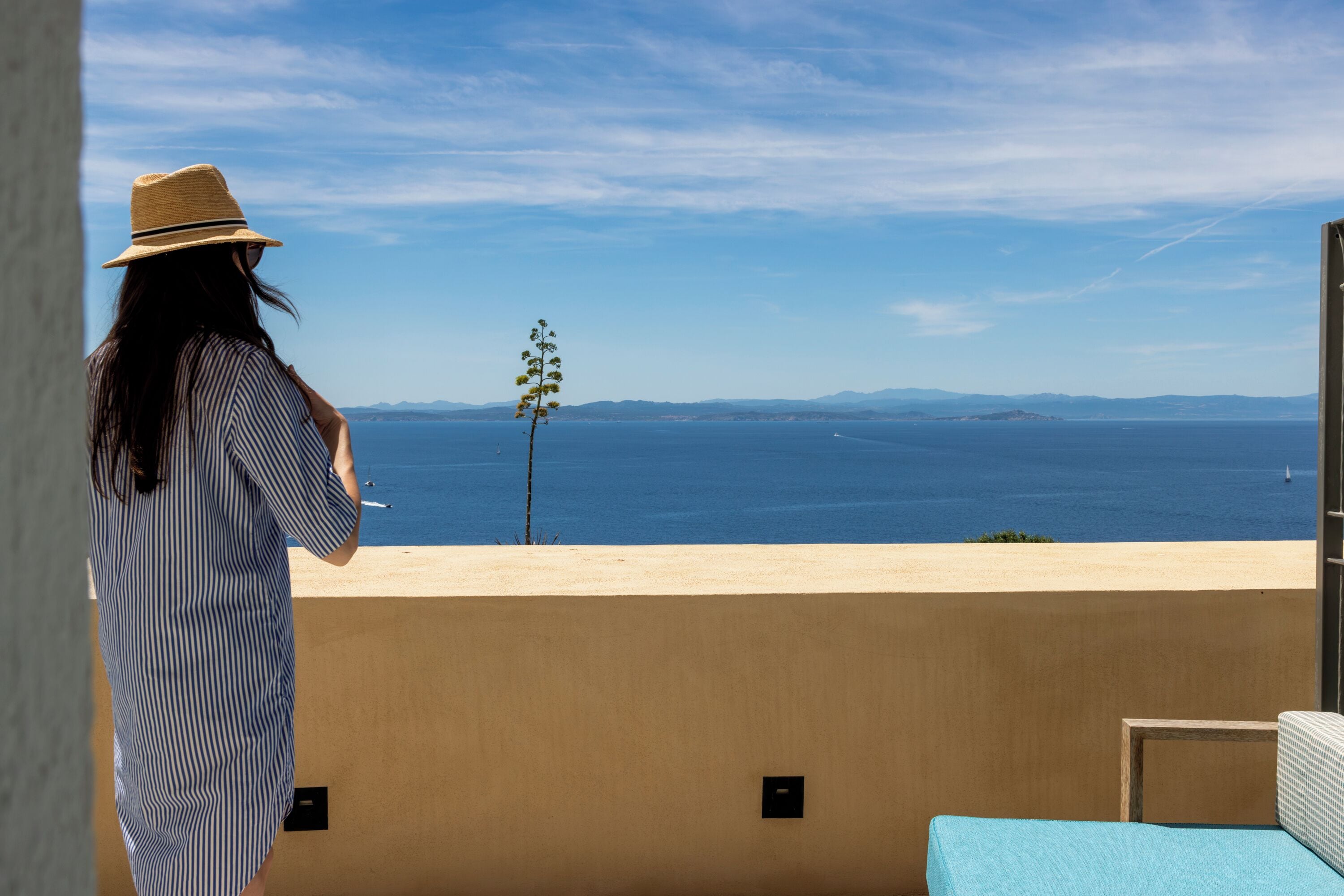 A person in a striped dress and hat standing on a balcony overlooking a body of water with distant landforms.