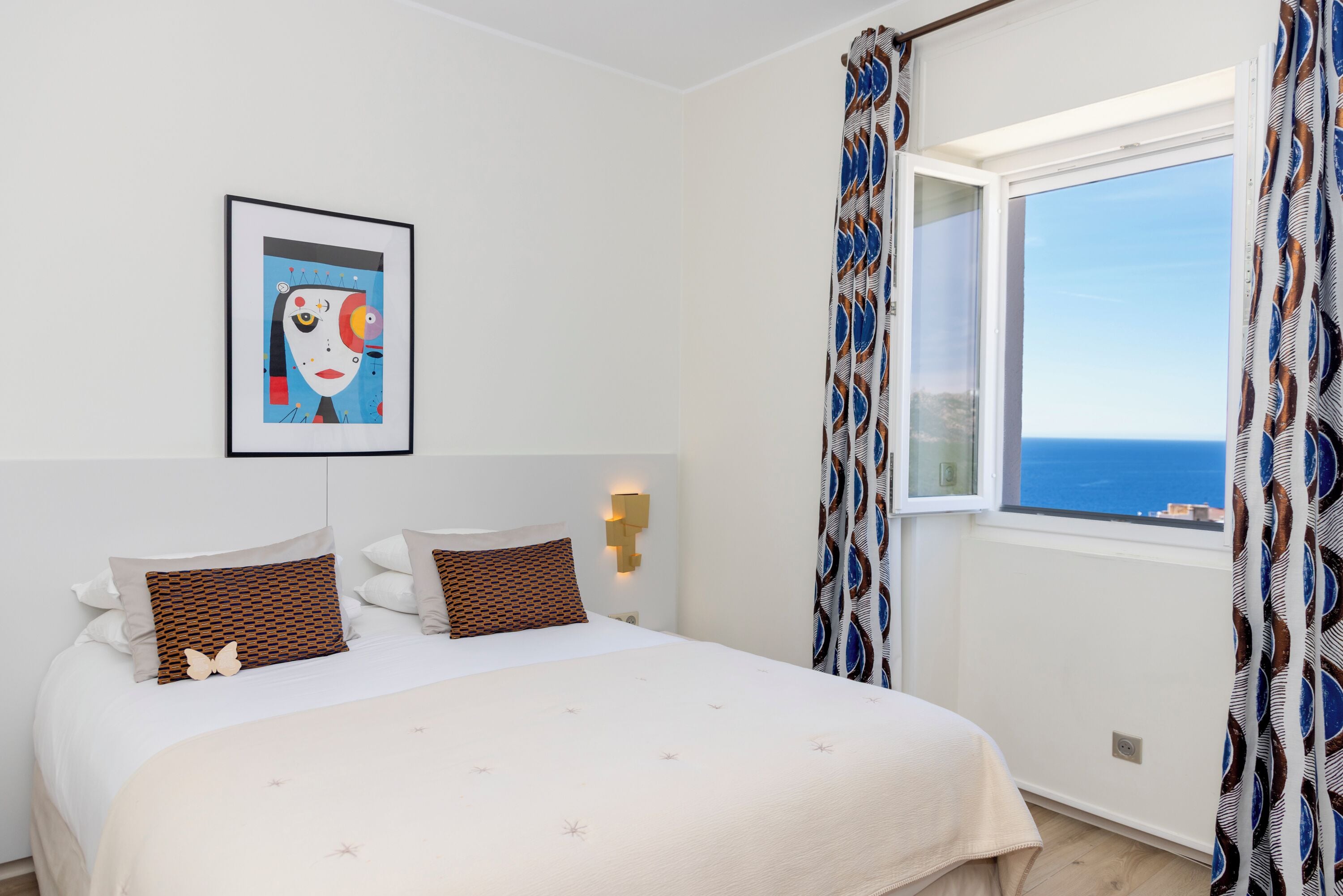 A bedroom with a bed, a framed artwork, a window with curtains, and a wall-mounted light.