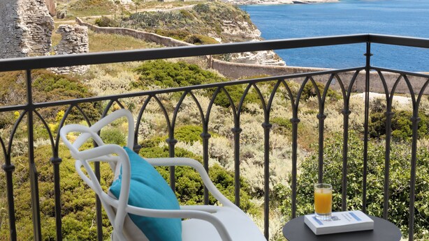 A balcony with a white chair and a table, overlooking a coastal landscape with cliffs and a historic building. - Hôtel Santateresa (Bonifacio)