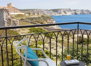 A balcony with a white chair and a table, overlooking a coastal landscape with cliffs and a historic building. - Hôtel Santateresa (Bonifacio)