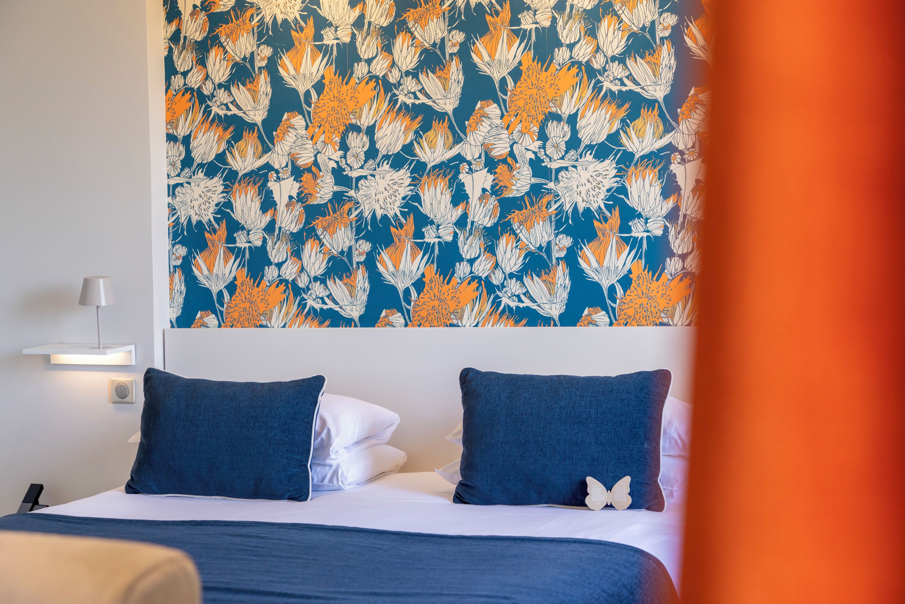 A hotel room with a bed, two pillows, a wall-mounted lamp, and a decorative wall hanging.