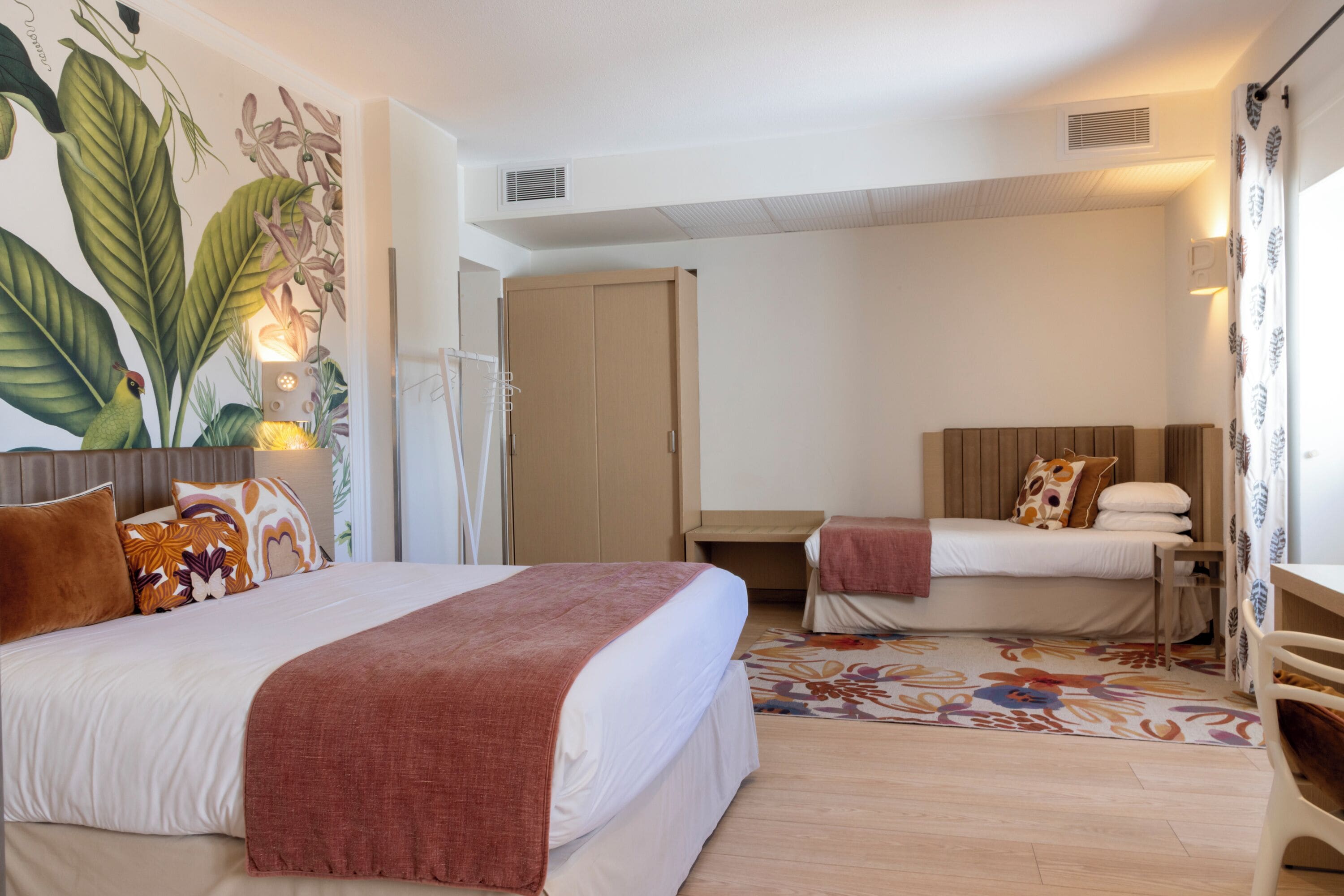 A hotel room with a large bed, a smaller bed, a desk, a wardrobe, and a wall mural.