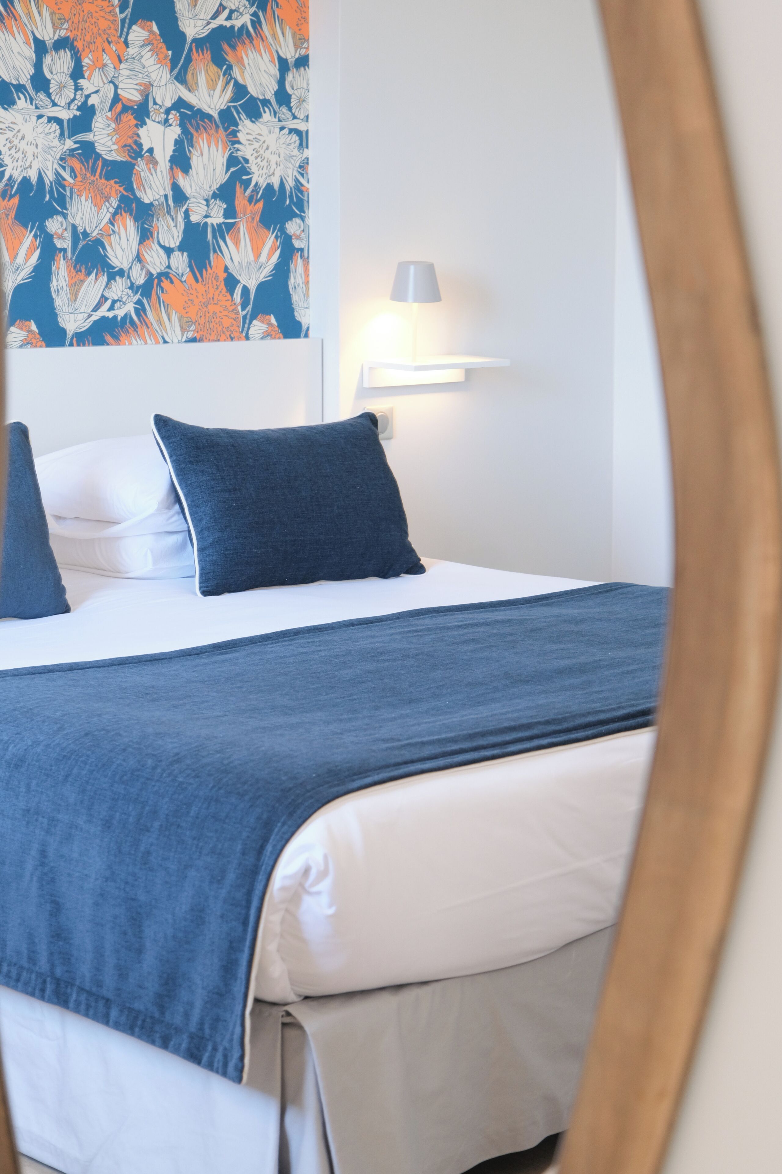 A neatly made bed with a blue blanket and pillows, a wall-mounted light, and a floral-patterned wall.