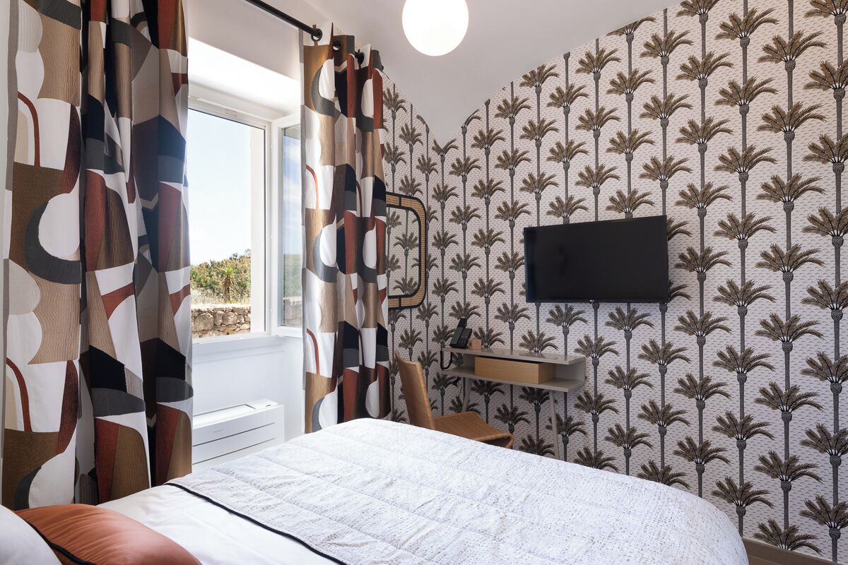 Een slaapkamer met een bed, een bureau, een stoel, een aan de muur bevestigde televisie en een raam met gordijnen.