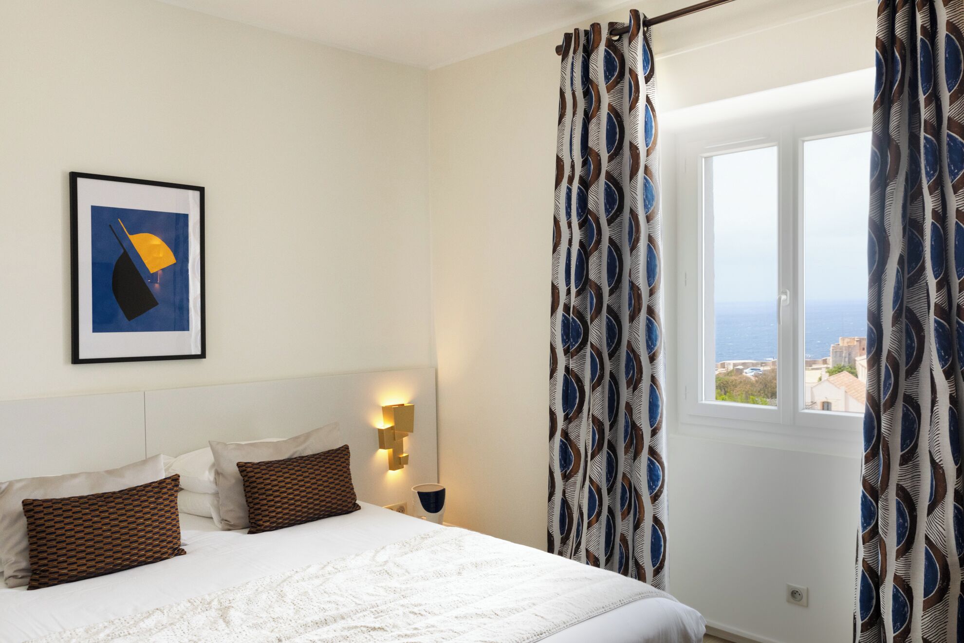 Une chambre à coucher avec un lit, des oreillers, une lampe fixée au mur, un tableau encadré et une fenêtre avec des rideaux.