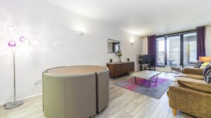A modern living room with a sofa, coffee table, and a TV. There is a dining table with chairs, a floor lamp, and a window with curtains. - Point West Apartments Gloucester Road (London)