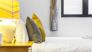 A neatly made bed with white bedding, a bedside table with a lamp, and a window with curtains. - Point West Apartments Gloucester Road (London)