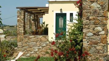 A stone-walled building with a green door, a pergola with chairs, and a garden with red flowers.